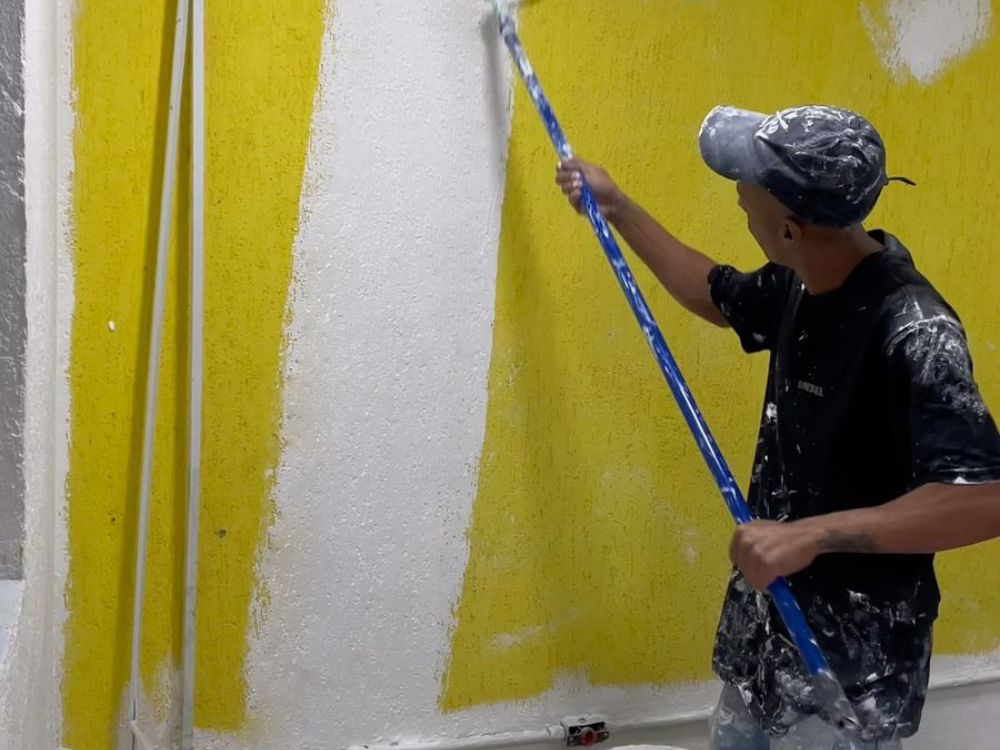 Pintor aplicando tinta branca com rolo sobre parede amarela, com balde da Will no chão